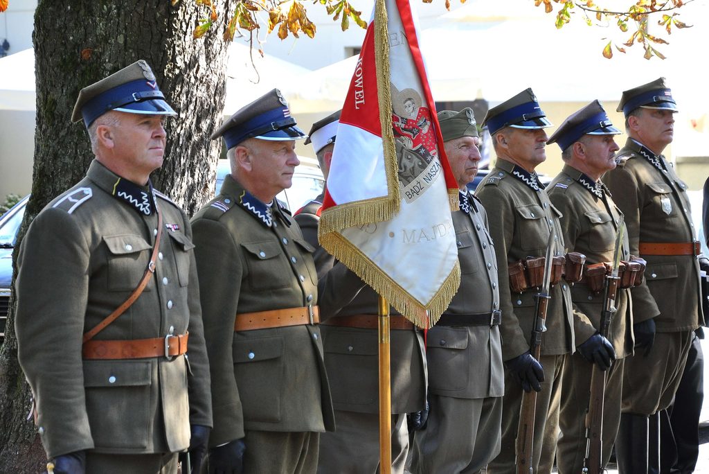Sześciu mężczyzn w mundurach wojskowych z granatowymi czapkami i skórzanymi pasami, trzymających szable, obok czerwono-biało-złotego sztandaru z haftowanym motywem.