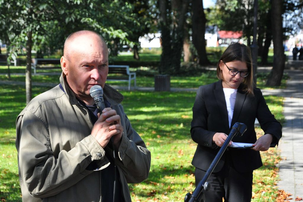 Mężczyzna z mikrofonem w beżowej kurtce i kobieta w czarnej marynarce z notatnikiem na tle zielonego parku z drzewami i ławkami.