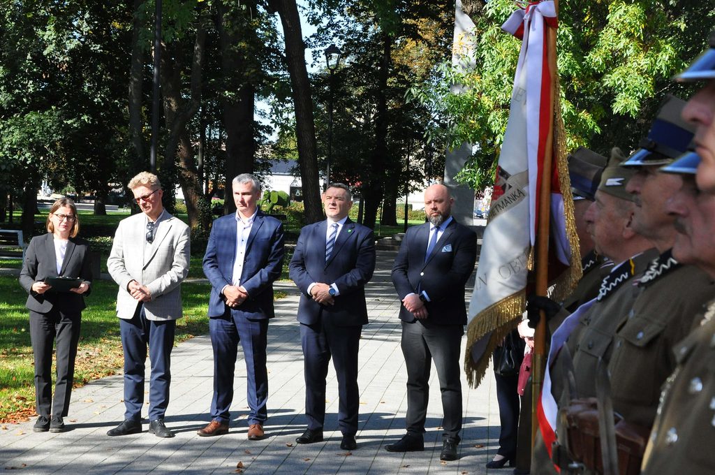 Grupa ludzi w formalnych ubraniach stoi na chodniku w parku, po prawej stronie żołnierze w mundurach i hełmach, trzymający flagę z biało-czerwonymi wstążkami.
