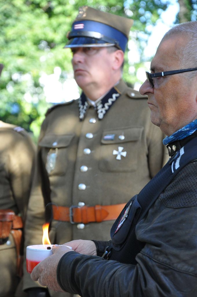 Starszy mężczyzna w czarnym stroju i okularach trzyma biało-czerwoną świecę, w tle mężczyzna w mundurze wojskowym z niebieską czapką i brązowym paskiem.