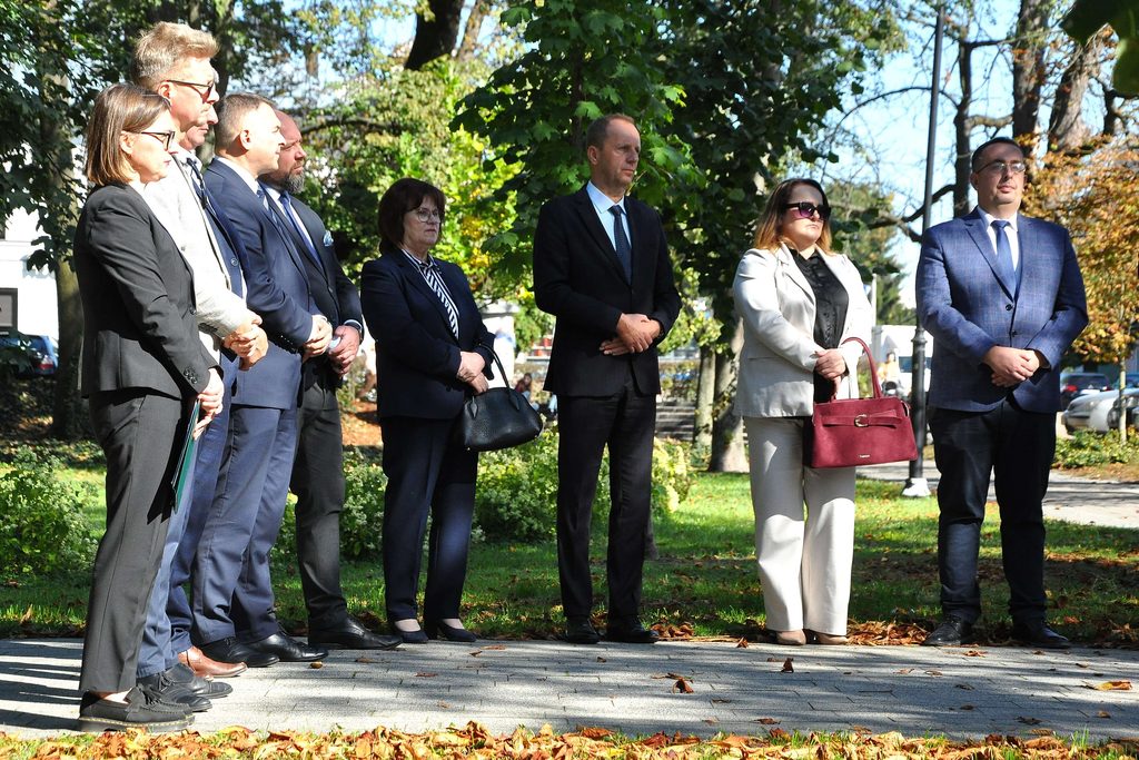 Siedem osób w garniturach i formalnych ubraniach stoi na chodniku na tle zielonych drzew i trawy, jesienne liście na ziemi, słoneczny dzień.