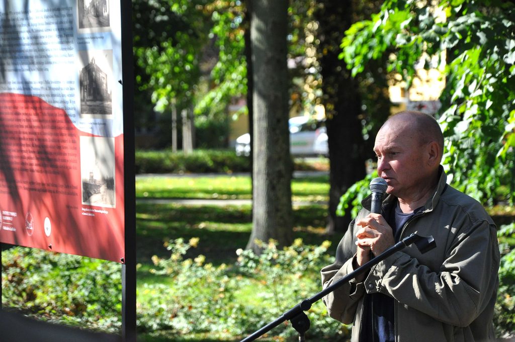 Mężczyzna w zielonej kurtce trzymający mikrofon, stojący na tle zielonych drzew i tablicy informacyjnej z tekstem i zdjęciami.