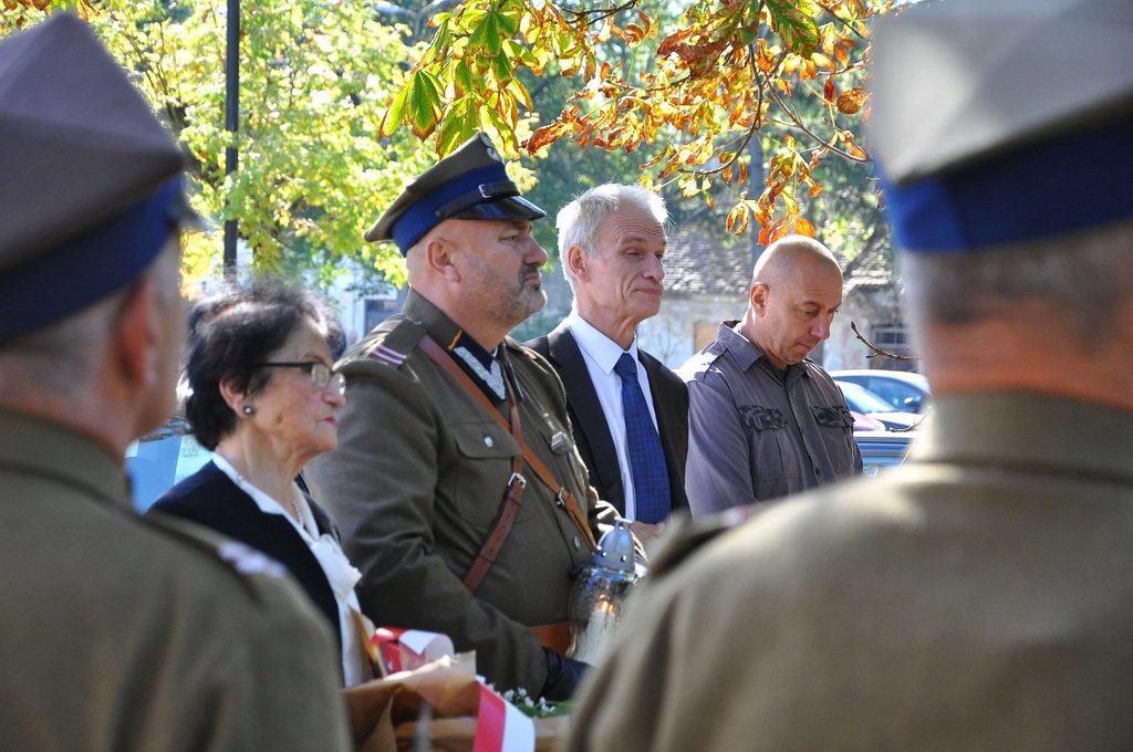 Grupa osób, w tym mężczyzna w mundurze wojskowym z niebieską czapką, kobieta w okularach oraz dwóch mężczyzn w garniturach, stoi na zewnątrz pod drzewem z jesiennymi liśćmi.