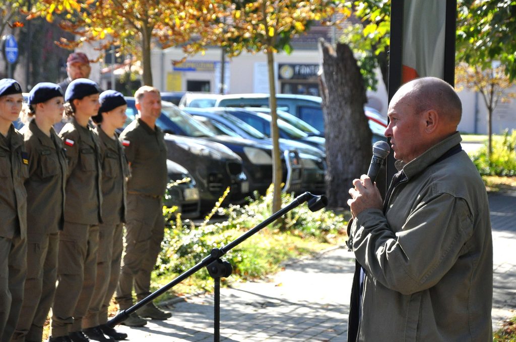 Mężczyzna z mikrofonem, w zielonej kurtce, przemawia do ustawionych w szeregu osób w mundurach z granatowymi beretami, w tle jesienne drzewa i zaparkowane samochody.