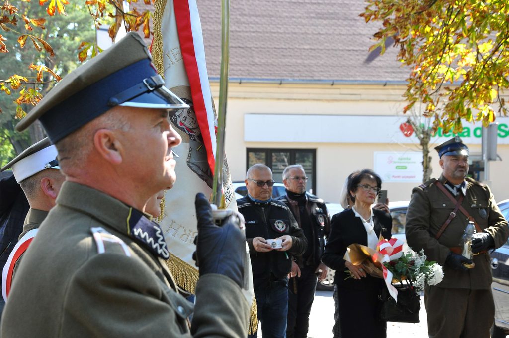 Mężczyzna w mundurze wojskowym trzymający flagę, w tle grupa osób, w tym kobieta z bukietem kwiatów, jesienne drzewa i budynek z oknami.