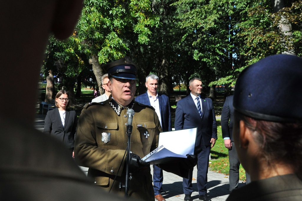 Mężczyzna w wojskowym mundurze z odznaczeniami mówi do mikrofonu, za nim stoją ludzie w garniturach i marynarkach, w tle zielone drzewa w parku.