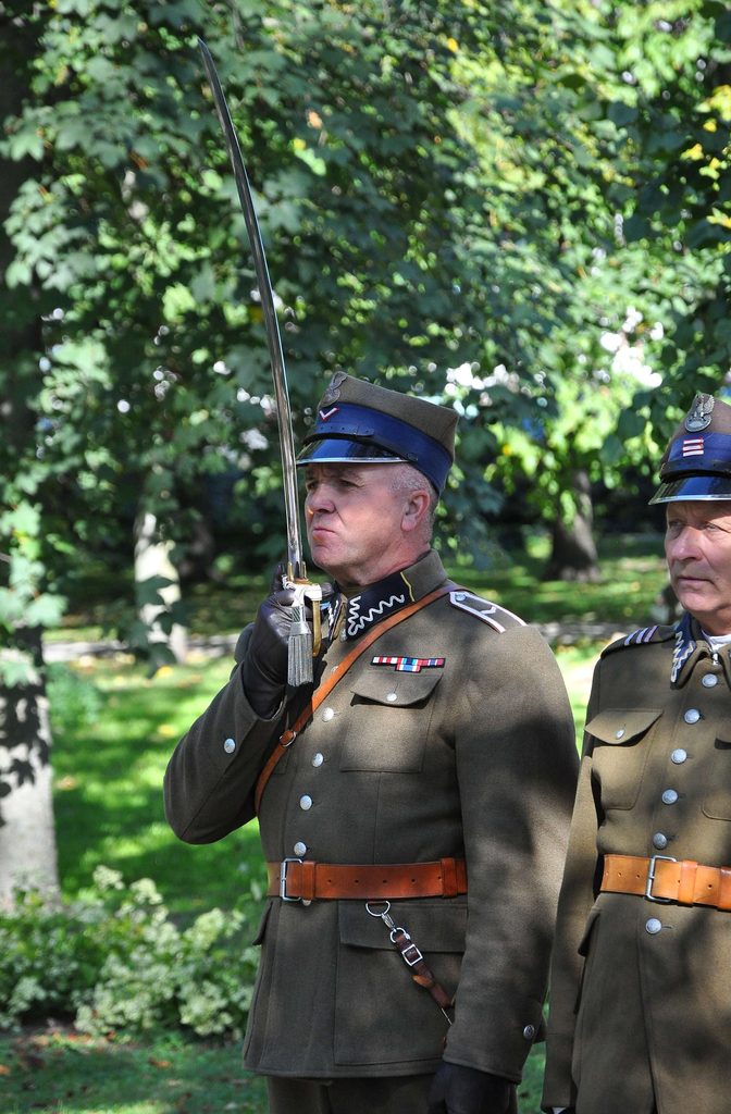 Dwóch mężczyzn w wojskowych mundurach z brązowymi pasami i granatowymi czapkami, jeden trzyma szablę pionowo, w tle zielone drzewa i trawa.