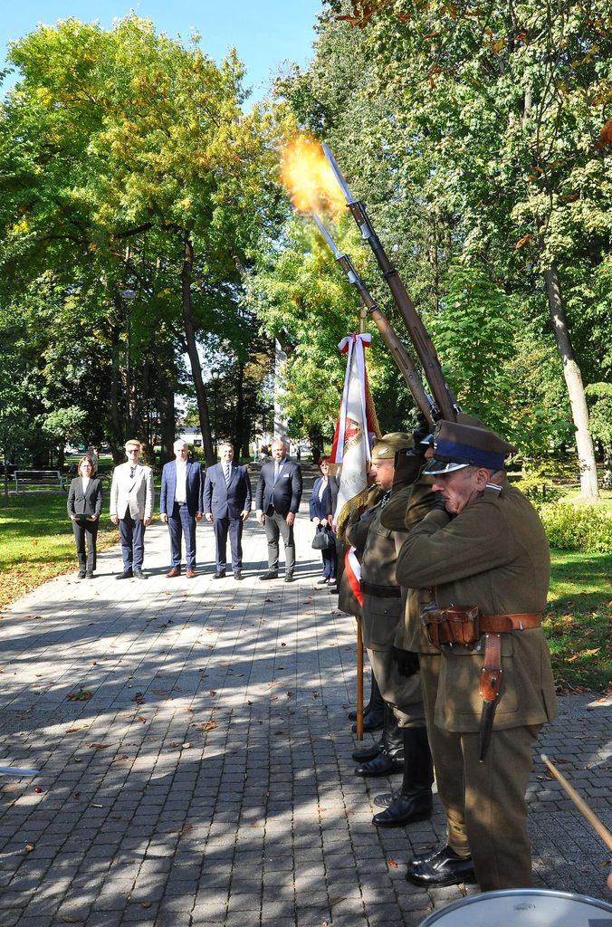 Żołnierze w mundurach historycznych z bronią z zamocowanymi bagnetami oddają salwę, w tle stoi grupa ludzi na tle zielonych drzew w parku.