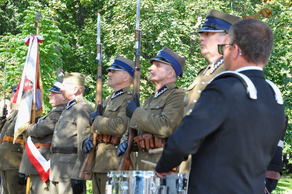 Grupa mężczyzn w mundurach wojskowych z granatowymi daszkami, trzymających broń z bagnetami, na tle zielonych drzew, obok mężczyzna w czarnym stroju z białymi szelkami.
