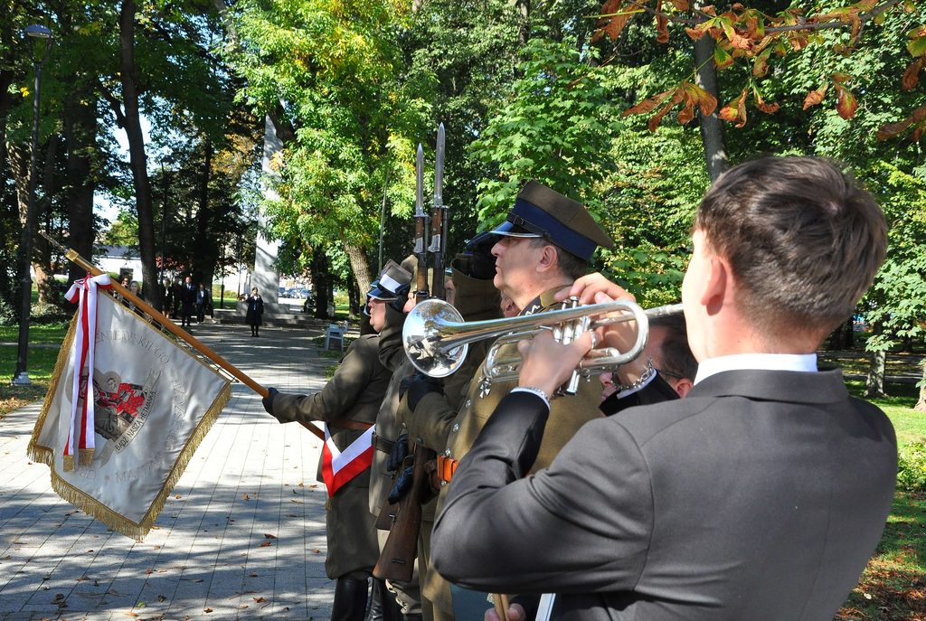 Mężczyzna grający na trąbce, obok żołnierze w mundurach z karabinami z bagnetami, jeden trzyma sztandar z białym i czerwonym orłem, park z zielonymi i brązowymi drzewami.