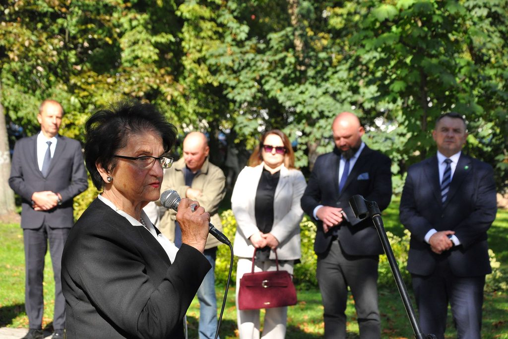 Kobieta w czarnym garniturze i okularach trzyma mikrofon, w tle pięć osób na tle zielonych drzew i trawy.