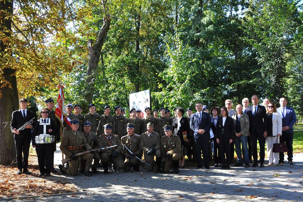 Grupa osób w mundurach wojskowych i formalnych ubraniach na tle drzew i zieleni w słoneczny dzień.