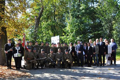 Grupa osób w mundurach wojskowych i formalnych ubraniach na tle drzew i zieleni w słoneczny dzień.